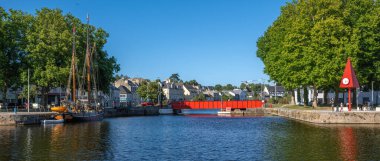 Caen, Fransa - 9 Ağustos 2025: Caen, Normandiya 'daki bir limanın manzaralı manzarası, kırmızı köprüsü, yanaşmış yelkenlileri ve açık mavi gökyüzünün altında suyla kaplı yemyeşil ağaçları.