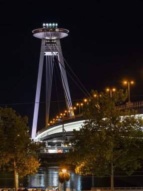 Bratislava, Slovakya - 18 Ekim 2025: Bratislava 'da sonbaharda renkli ağaçlarla çevrili UFO şeklinde bir gözlem güvertesi bulunan modern bir asma köprünün gece görüntüsü.