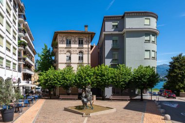 Lugano, İsviçre - 9 Temmuz 2025: Lugano 'da ağaçlar ve binalarla çevrili, arka planda açık mavi gökyüzü bulunan bir çeşme ve heykel manzaralı bir meydan