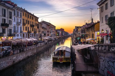 Milan, İtalya - 9 Aralık 2025: Gün batımında Milano 'da Naviglio Büyük Kanalı' nın manzaralı manzarası, renkli binalar su üzerinde yüzüyor, insanlar gezinti güvertesi boyunca yürüyor, sudaki yansımalar...