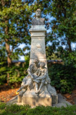 Nantes, Fransa - 5 Ağustos 2025: Jules Verne 'in üzerinde büst bulunan taş bir heykel ve üste bulanık ağaçlarla çevrili iki kişinin okuduğu bir heykel.