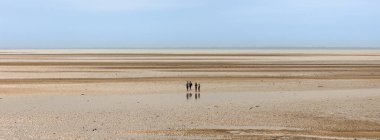 Normandiya 'da Atlantik Okyanusu' nun engin, boş bir sahilinde yürüyen dört kişi, alçak gelgitte, bulutlu bir gökyüzü, minimalizm.