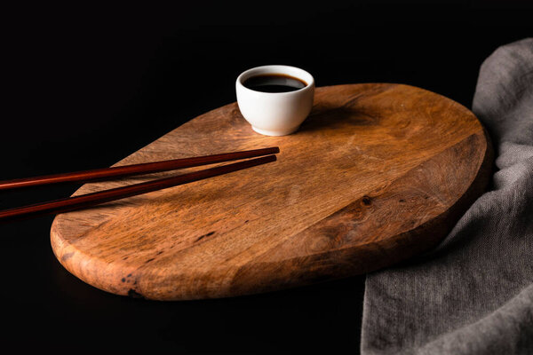 Two Wooden Sushi Chopsticks, Bamboo Wood Kitchen Board, White Soy Sauce Gravy Boat And Grey Napkin. Japan Or China Cuisine Still Life On Dark Black Background.