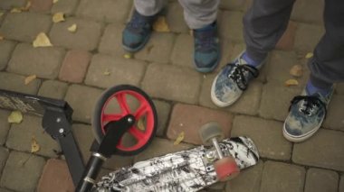 İki genç bir peni tahtası ve kepçeyle çevrili. Çerçevede sadece tekerleğin döndüğü bacaklar ve scooter var. Parkta sonbahar günü, yeşil-sarı yapraklar. yavaş çekim.