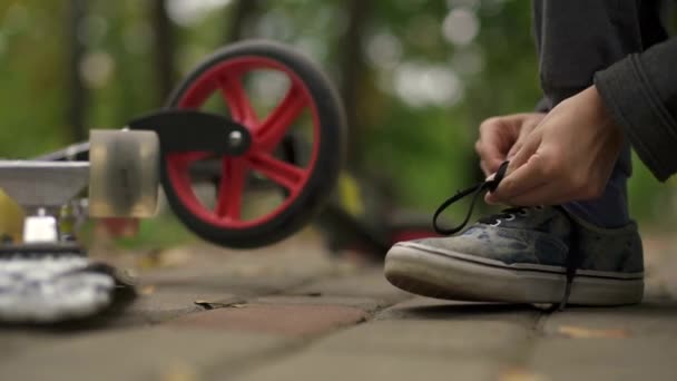 Хлопець прив'язав шнурівку до спини скутера. В кадрі лише ноги і скутер, на яких крутиться колесо. Осінній день у парку, зелено-жовте листя. повільний рух — стокове відео