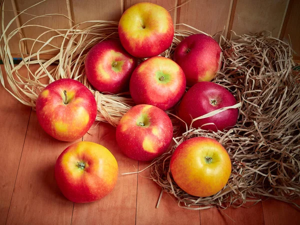 Bright red and yellow apples are stored in hay on a wooden surface