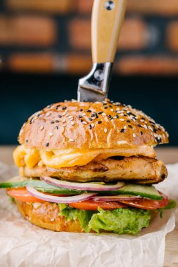 Brioche Bun 'dan leziz sulu bir burger. Tavuk biftek, peynir, soğan, domates, salatalık, marul, saplanmış bıçak, sos. Kafe masası. Tahta.