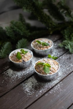 Koyu ahşap masa ve köknar dalının arkasında duran naneli yapraklar ve pudra şekerli üç kekin dikey görüntüsü