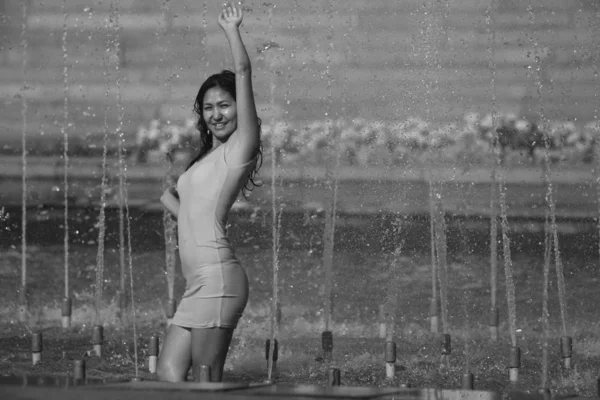 Chica en un vestido elegante con el pelo largo en gotitas de agua en la fuente de la ciudad —  Fotos de Stock