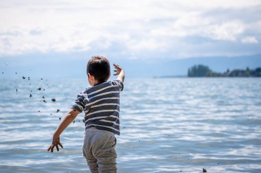 Child playing sands head down on the beach. One little Asian boy in casual clothing beside water in Switzerland. Happy childhood.