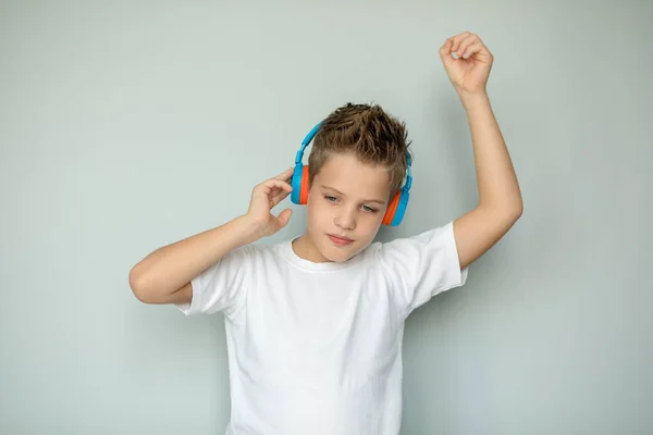 A boy in a white T-shirt listens to music with headphones. The teenager moves to the beat of the music, isolated on a gray background. Large portrait.