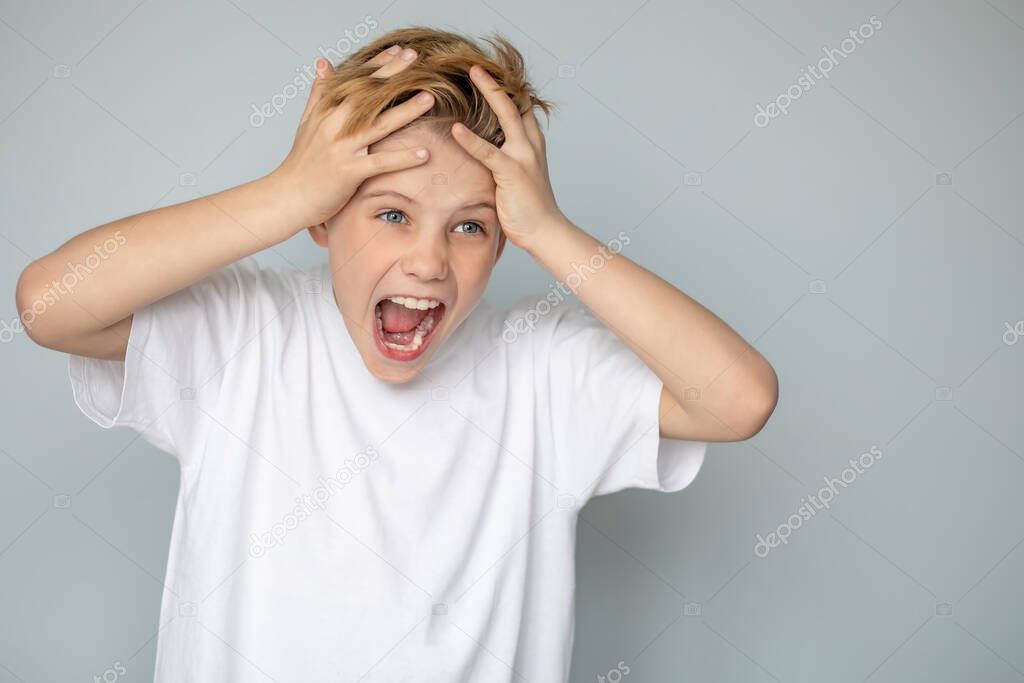 A teenage boy in a casual T-shirt shouts, holding his head with his hands, with an aggressive expression on his face. Youth problems, adolescence.