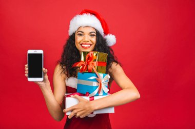 Kırmızı Noel Baba kıyafetli, kırmızı arka planda cep telefonu olan mutlu Afrikalı Amerikalı kadın. Hediye kutuları tutuluyor. 