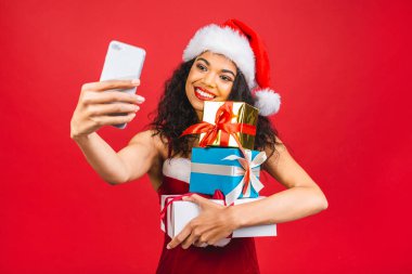 Kırmızı Noel Baba kıyafetli, kırmızı arka planda cep telefonu olan mutlu Afrikalı Amerikalı kadın. Hediye kutuları tutuluyor. 