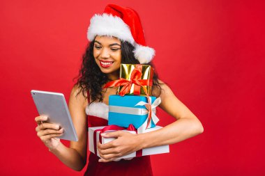 Güzel gülümseyen mutlu Afro-Amerikan siyah kadın Noel Baba kıyafetleri içinde kırmızı arka planda izole edilmiş. Yeni yıl ve Noel konsepti. Hediye kutusu tutuluyor ve tablet kullanılıyor. 