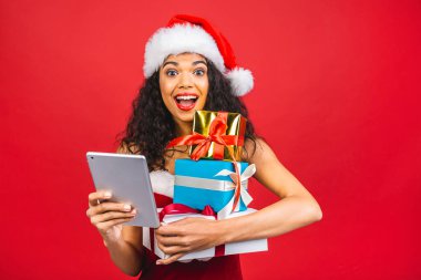 Güzel gülümseyen mutlu Afro-Amerikan siyah kadın Noel Baba kıyafetleri içinde kırmızı arka planda izole edilmiş. Yeni yıl ve Noel konsepti. Hediye kutusu tutuluyor ve tablet kullanılıyor. 
