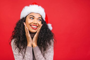 Kırmızı arka planda izole edilmiş Noel Baba şapkası giyen güzel Afro-Amerikan kadın modelin portresi.. 