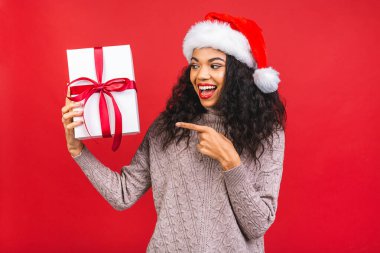 Güzel gülümseyen mutlu Afro-Amerikan siyah kadın Noel Baba kıyafetleri içinde kırmızı arka planda izole edilmiş. Yeni yıl ve Noel konsepti. Hediye kutusu tutuluyor. 
