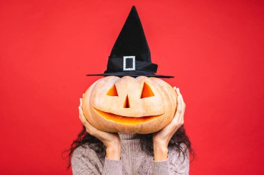 Cadılar Bayramı Afro-Amerikan cadı kadın portresi kırmızı arka planda saklanmış bir balkabağı ve büyük bir şapka tutuyor. Cadılar Bayramı tarzı, Cadılar Bayramı partisi.