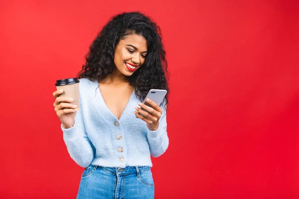 Happy young african american woman casually dressed standing isolated over pink background, holding mobile phone and cup of coffee or tea.