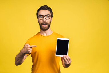 Ürün sunumu. Terfi ettim. Genç sakallı adam elinde tablet bilgisayarla boş ekranı tutuyor. Sarı arkaplan üzerinde izole edilmiş.