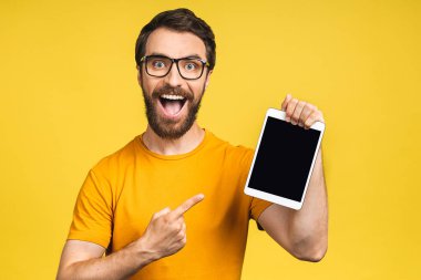 Ürün sunumu. Terfi ettim. Genç sakallı adam elinde tablet bilgisayarla boş ekranı tutuyor. Sarı arkaplan üzerinde izole edilmiş.