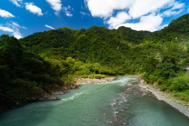 Açık mavi-yeşil bir nehir, bulutlarla dolu parlak bir gökyüzünün altında orman dağlarının arasında dolanıyor..