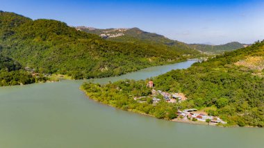 Yoğun ormanlık tepeler vadisinde kıvrılan geniş bir zümrüt nehri, berrak mavi bir gökyüzünün altında kıyı şeridi boyunca yerleşmiş dağınık kırsal evler..