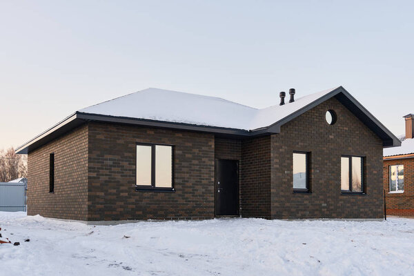 Одноэтажный кирпичный дом на фоне снежного зимнего вечера. Строительство малоэтажных зданий для дальнейшей продажи. Концепция строительного бизнеса.