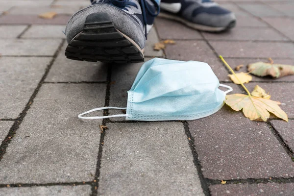A man in blue sneakers steps on a protective mask on the ground. Medium shot, male legs and mask. Concept, stop coronavirus.
