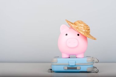 Hasır şapkalı bir kumbaranın yanında minimalist bir arka planda mavi bir bavul. Seyahat, tatil tasarrufu ve mali planlama kavramı.