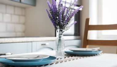 Masa örtüsünde beyaz tabaklar, çatallar ve mavi şarj aletleri olan yemek masası. Ortasında mor çiçekler olan cam vazo, arka planda mutfak, kapalı alanda doğal ışık..