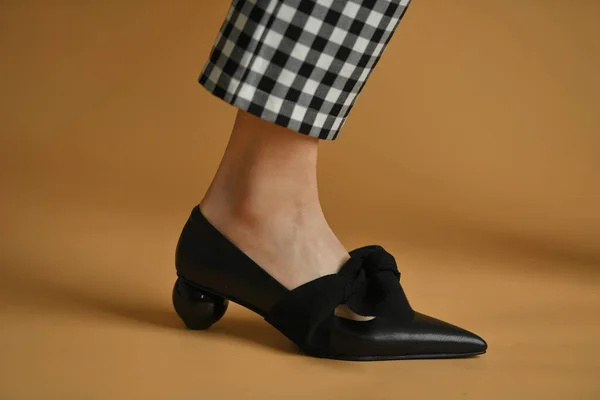 Stylish female shoe with sharp sock, unique sphere heel and a cloth bow on top. Beautiful leg ankle and plaid pants trews. Natural colors and shadow, isolated on brown background. Fashion concept.