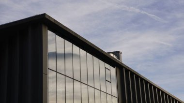 Camdan cepheli modern bir bina bulutlu gökyüzünü yansıtıyor, temiz hatları ve minimalist bir tasarımı var.