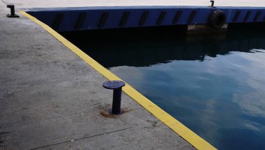 Beton iskele kenarında sarı bir güvenlik şeridi, ön planda mavi bir demirleme bölmesi ve mavi rıhtım duvarının yanında gökyüzünü yansıtan sakin bir liman suyu..