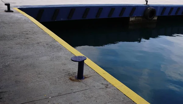 Beton iskele kenarında sarı bir güvenlik şeridi, ön planda mavi bir demirleme bölmesi ve mavi rıhtım duvarının yanında gökyüzünü yansıtan sakin bir liman suyu..