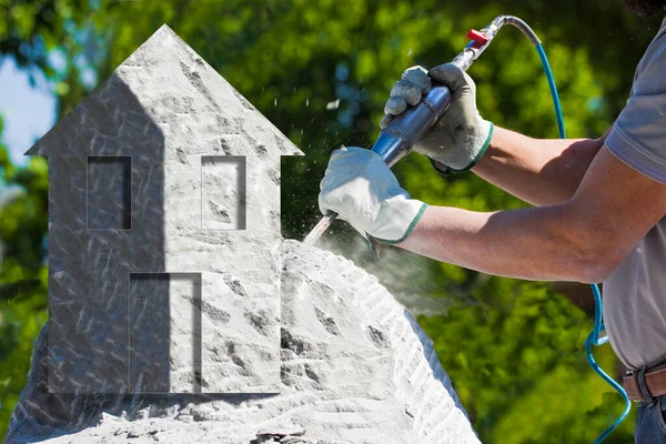 Man at work with compressed air chisel and protective gloves carving a little house on a stone block - concept image - It is not a real image it is a photomontage.