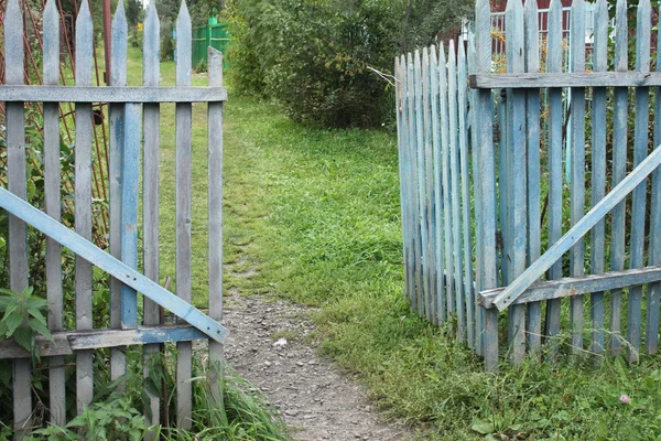 Een blauwe oude haveloze poort naar de tuin van een datsja in een dorp met grond en gras en bomen Stockafbeelding