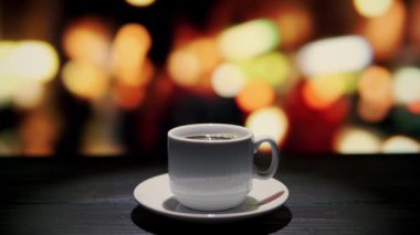 Gece şehrine bakan pencerenin önündeki bir kafede, ahşap masanın üzerinde bir fincan kahve.