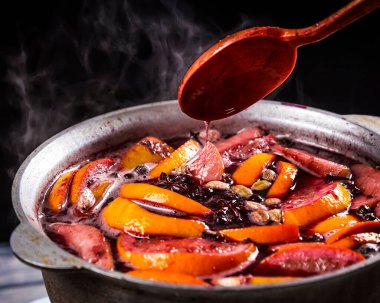 Büyük çaydanlık dolusu geleneksel sıcak buharlı Noel içeceği kaynatılmış kırmızı baharatlı şarap elma ve siyah arka planda tahta kaşıkla narenciye. Noel partisi.