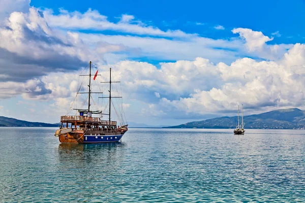Vintage mast wooden sailing ship for sea tours in Saranda gulf, Albania with red state albanian flag with black double-headed eagle. Small yacht and Corfu island on the horizon