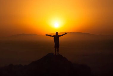 Dağda gün batımını kolları açık izleyen bir adamın silueti.