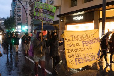 Kadınlar, kadın hakları lehinde bir protesto posteri tutuyor. Yağmur yağarken ortaya çıkan bir tezahür.