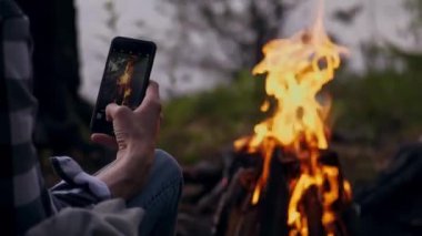 Bir adam akıllı telefondaki şenlik ateşini fotoğraflıyor. Kamp yapmak. Dağlarda yürüyüş. Turizm. Seyahat