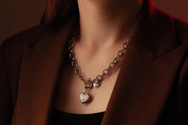Przycięty z bliska portret młodej kobiety z idealną jedwabistą skórą, demonstrujący srebrny łańcuszek biżuterii z medalionem na szyi w brązowej kurtce — Zdjęcie stockowe