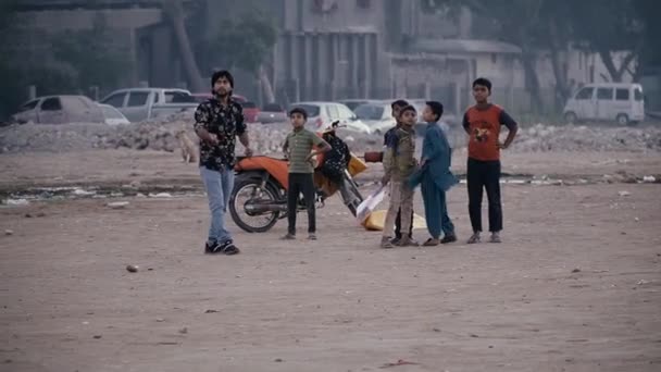 Karachi, Paquistão - 24 de fevereiro de 2021: Adolescentes muçulmanos de pele escura em roupas sujas e baratas ficam ao lado de uma motocicleta em um terreno baldio em um bairro pobre, assistindo seu amigo mais velho voar um papagaio — Vídeo de Stock