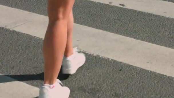 Una hermosa chica delgada en pantalones cortos de color turquesa y zapatillas blancas con una marcha rápida y segura cruza la carretera a lo largo del cruce peatonal en verano. Camino empedrado en la ciudad. Filmación 4K — Vídeo de stock