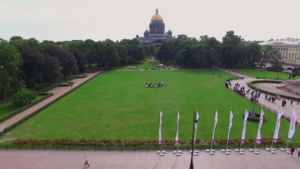 Lidé procházet po zeleném trávníku v blízkosti katedrály sv. Izáka v centru Petrohradu, Rusko. Quadcopter letí zpět a objevují se turisté, památky, auta, řeka Neva a lodě. 4K Aerial — Stock video