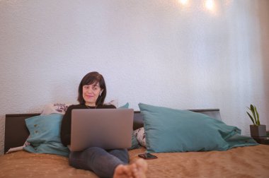Yatak odasında dizüstü bilgisayarda çalışan gülümseyen genç bir kadın. Evden bilgisayarda çalışma kavramı. Evden sosyal uzaklık.