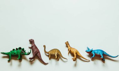 Beş oyuncak dinozor, beyaz bir arkaplana karşı bir sırada duruyor..